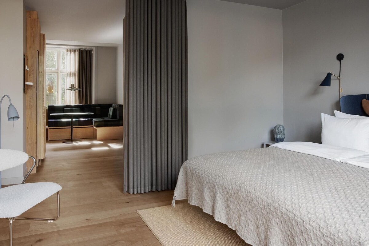 A modern bedroom with a large bed, a seating area with a sofa and chairs, a small table, and a lamp.