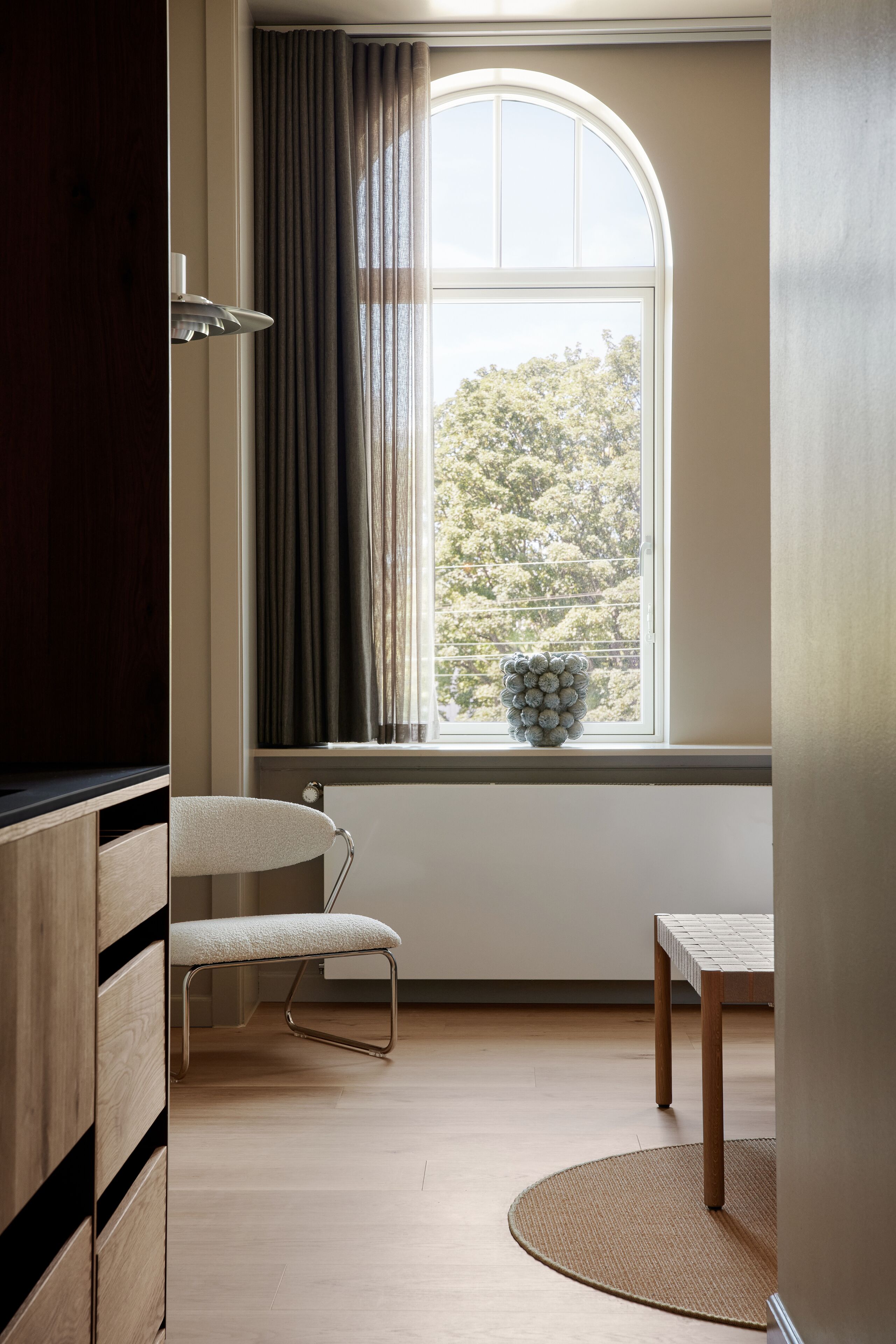 A room with a large arched window, a wooden floor, a white chair, a small round table, and a wall-mounted lamp.