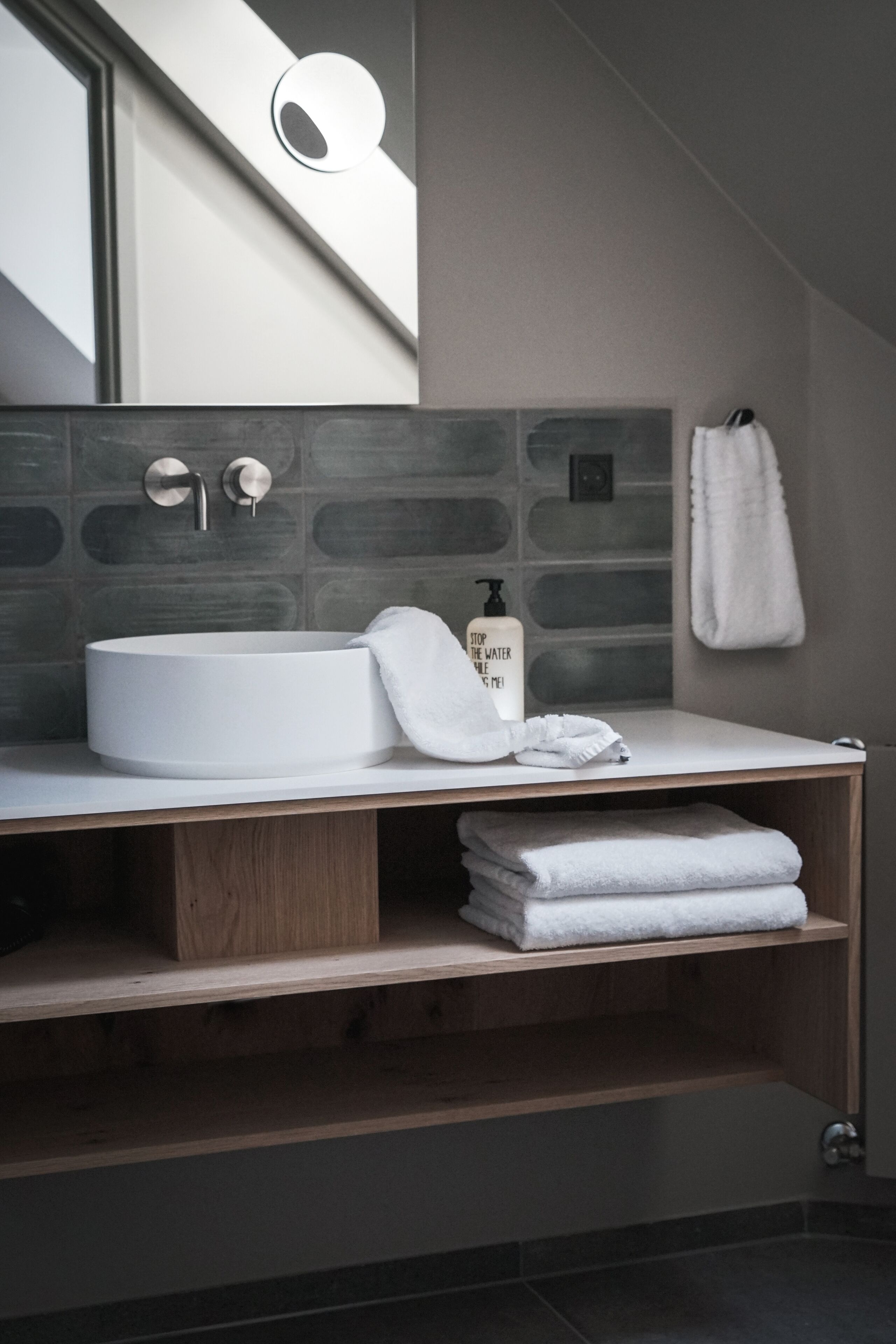 A modern bathroom with a round sink, wooden shelves, a towel rack, and a mirror.