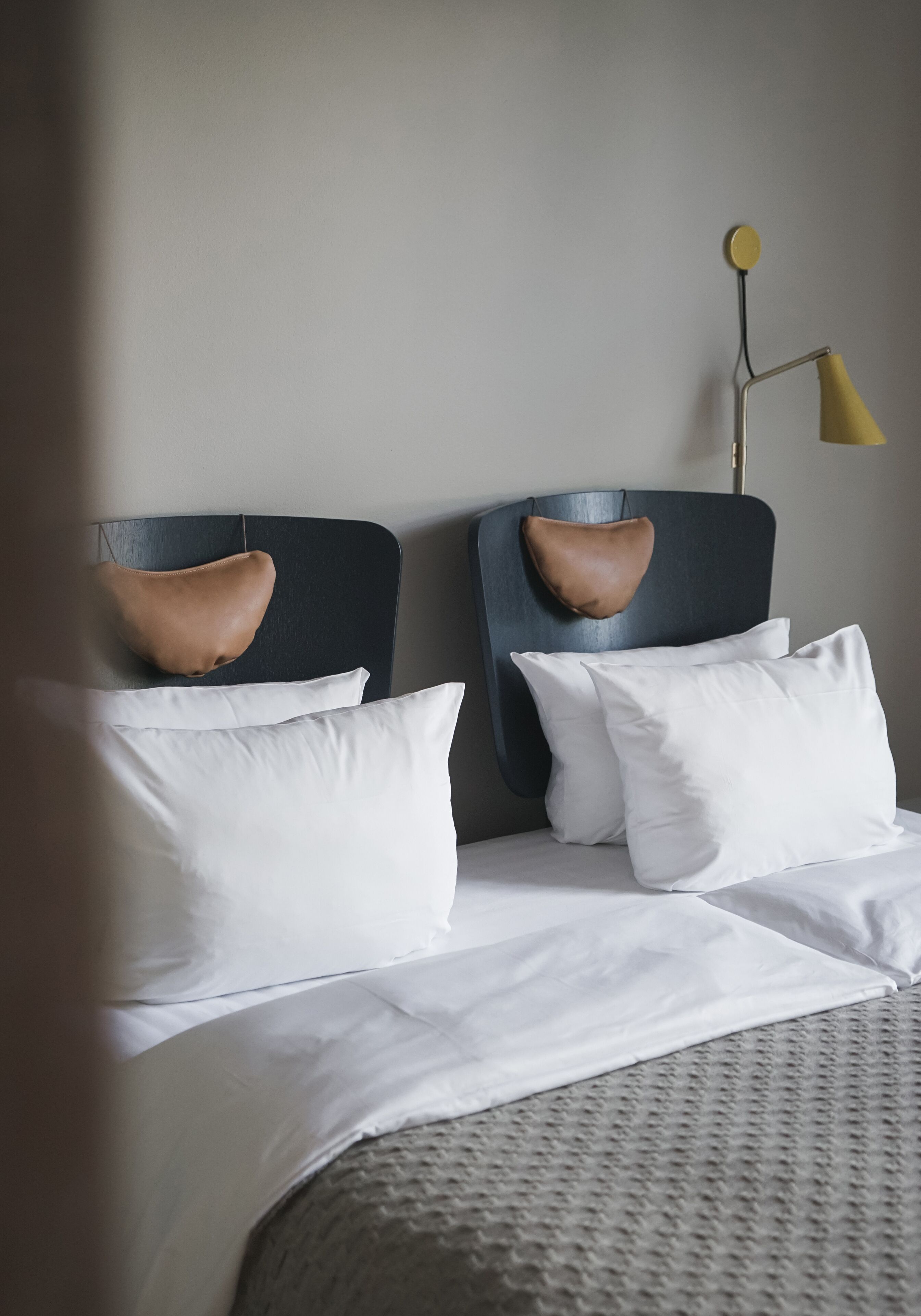 A neatly made bed with white linens and two pillows, a modern lamp with a yellow shade, and a headboard with a leather accent.