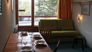 A dining area with a wooden table set for four, a green sofa, and a small stool. A window with curtains is visible, and a picture frame hangs on the wall. - 10m from ski slopes, 10th floor, balcony, tv, 28m², Les Arcs (Les Arcs)