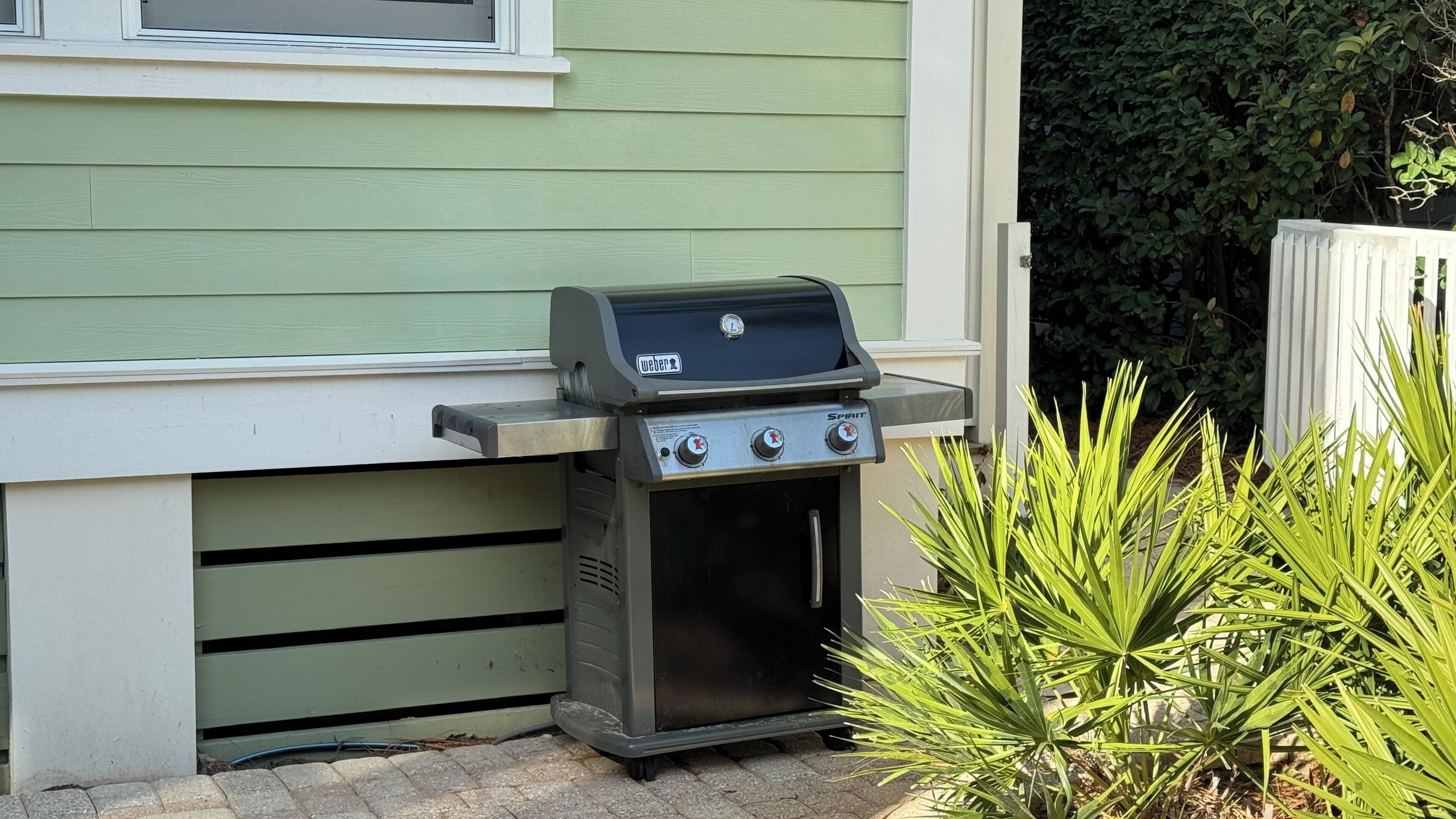 A Weber Spirit portable grill with a built-in oven, positioned against a green house exterior.