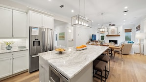 A modern kitchen with stainless steel appliances, a marble island, and a dining area with wooden flooring and a fireplace. - All Decked Out (Santa Rosa Beach)