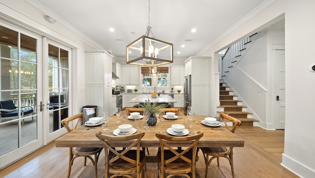 A spacious dining area with a large wooden table set for six, surrounded by wooden chairs. A modern chandelier hangs above, and the room features white cabinetry and a visible kitchen area in the background. - All Decked Out (Santa Rosa Beach)