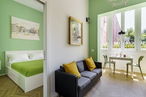 Una habitación con cama, sofá, mesa y sillas. Se ve una ventana con vistas a edificios.