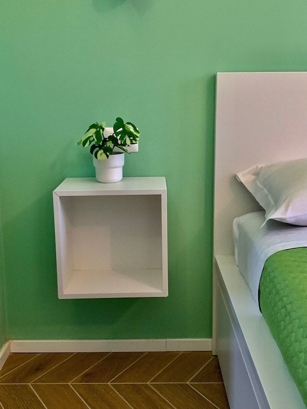 Un dormitorio con pared verde, una mesita de noche blanca con planta en maceta y una cama con ropa de cama blanca y almohada verde.