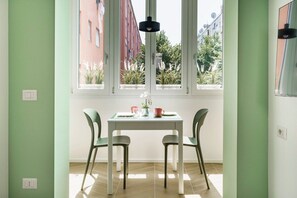 Zona pranzo con tavolo e sedie, una finestra vista edifici e vegetazione, e un lampadario.