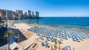 Ein Strand mit blauen Sonnenschirmen und einer Promenade gesäumt von Palmen.