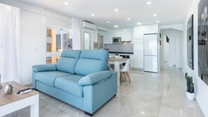 Ein modernes Wohnzimmer mit einem blauen Sofa, einem Esstisch mit Stühlen und einer Küchenzeile mit weißen Schränken und einer Mikrowelle.