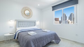 Ein modernes Schlafzimmer mit einem großen Bett, einem runden Spiegel und Blick auf die Stadt durch ein Fenster.