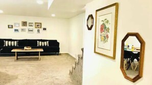 A living room with a black sofa, a wooden coffee table, and a carpeted floor. There are framed pictures on the wall and a mirror.
