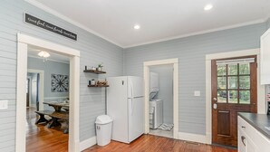 Une cuisine avec un plancher en bois, un réfrigérateur blanc et un four à micro-ondes. Une porte donne accès à une salle de bain, et un panneau est accroché au mur avec l’inscription suivante : « Bonne nourriture. Bons amis. Bons moments. »