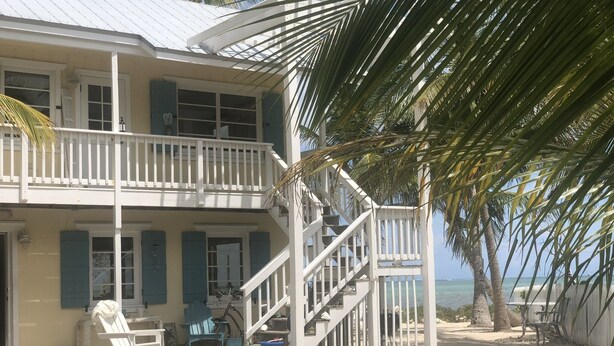 A two-story beachfront house with a balcony, a staircase, and a sandy beach in the background.