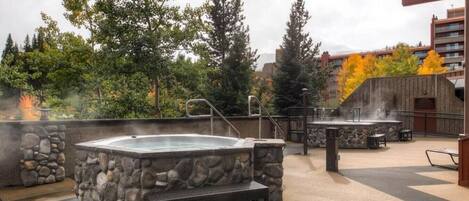 Un espace extérieur aménagé avec un jacuzzi, des murs en pierre et un banc, le tout entouré d’arbres et avec un bâtiment visible au loin.