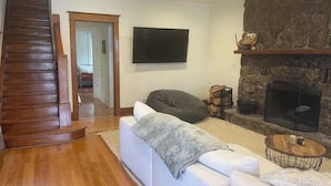 A living room with a white sofa, a stone fireplace, a flat-screen TV, and a wooden staircase.