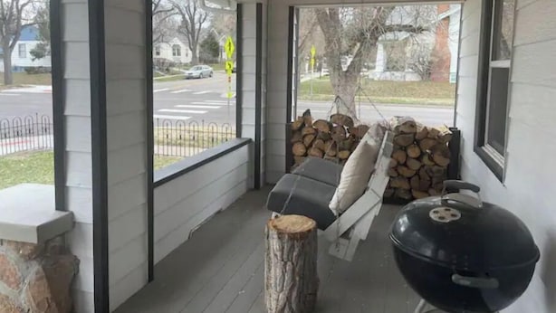 A porch with a black grill, a stack of firewood, and a bench with pillows.