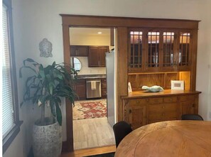 A dining area with a wooden table and chairs, a potted plant, and a view into the kitchen area with wooden cabinets and a refrigerator.
