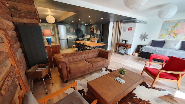 Ein gemütliches Wohnzimmer mit einem braunen Sofa, einem Holztisch und einem roten Sessel. Im Hintergrund ist eine Küchenzeile mit modernen Geräten und einem Esstisch zu sehen.