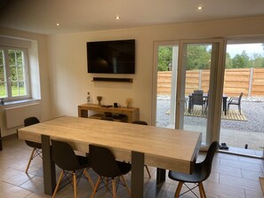 Un espace repas moderne avec une table en bois et des chaises noires, un téléviseur à écran plat fixé au mur au-dessus, et une porte-fenêtre coulissante donnant sur une terrasse extérieure.