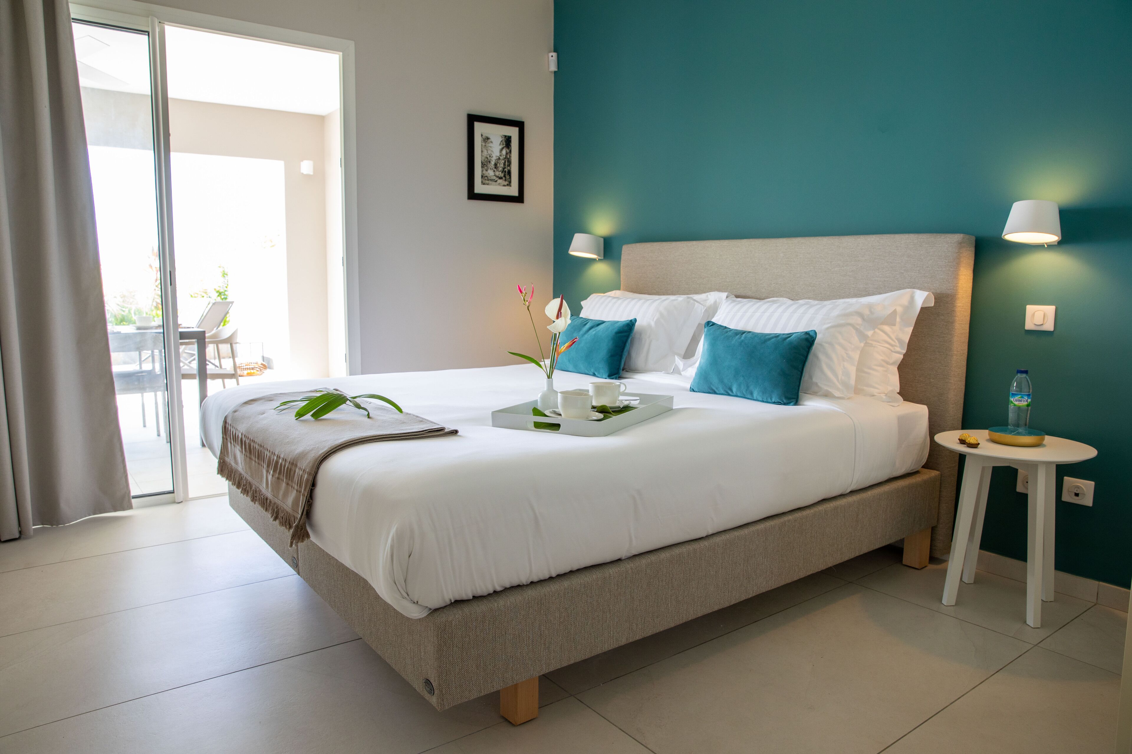 A neatly made bed with white and blue bedding, a bedside table with a lamp, and a view of the outdoors through a sliding door.