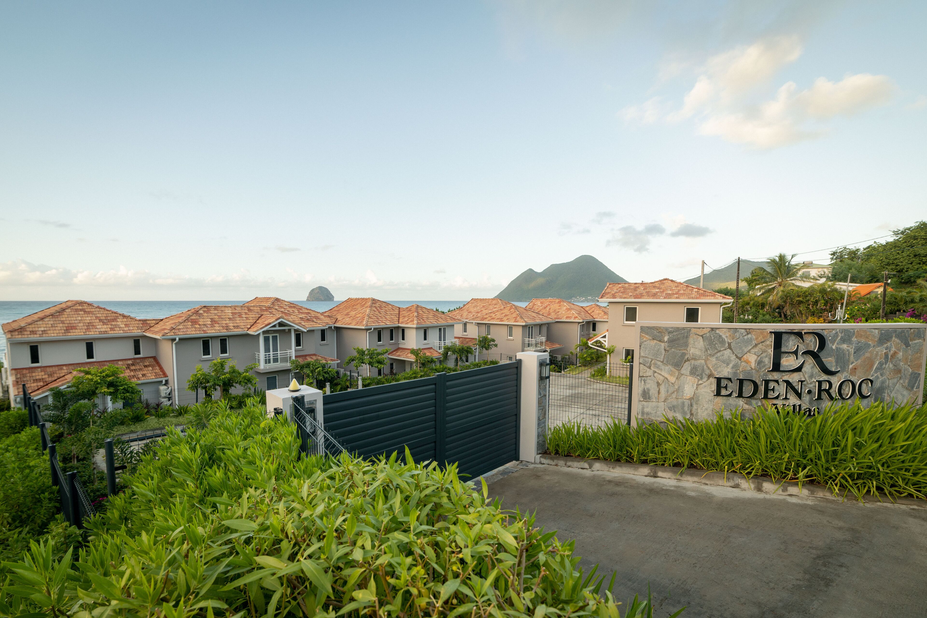 Un lotissement résidentiel fermé, avec des maisons, un panneau en pierre portant l’inscription « EDEN ROC », et une vue sur la mer et les montagnes en arrière-plan.