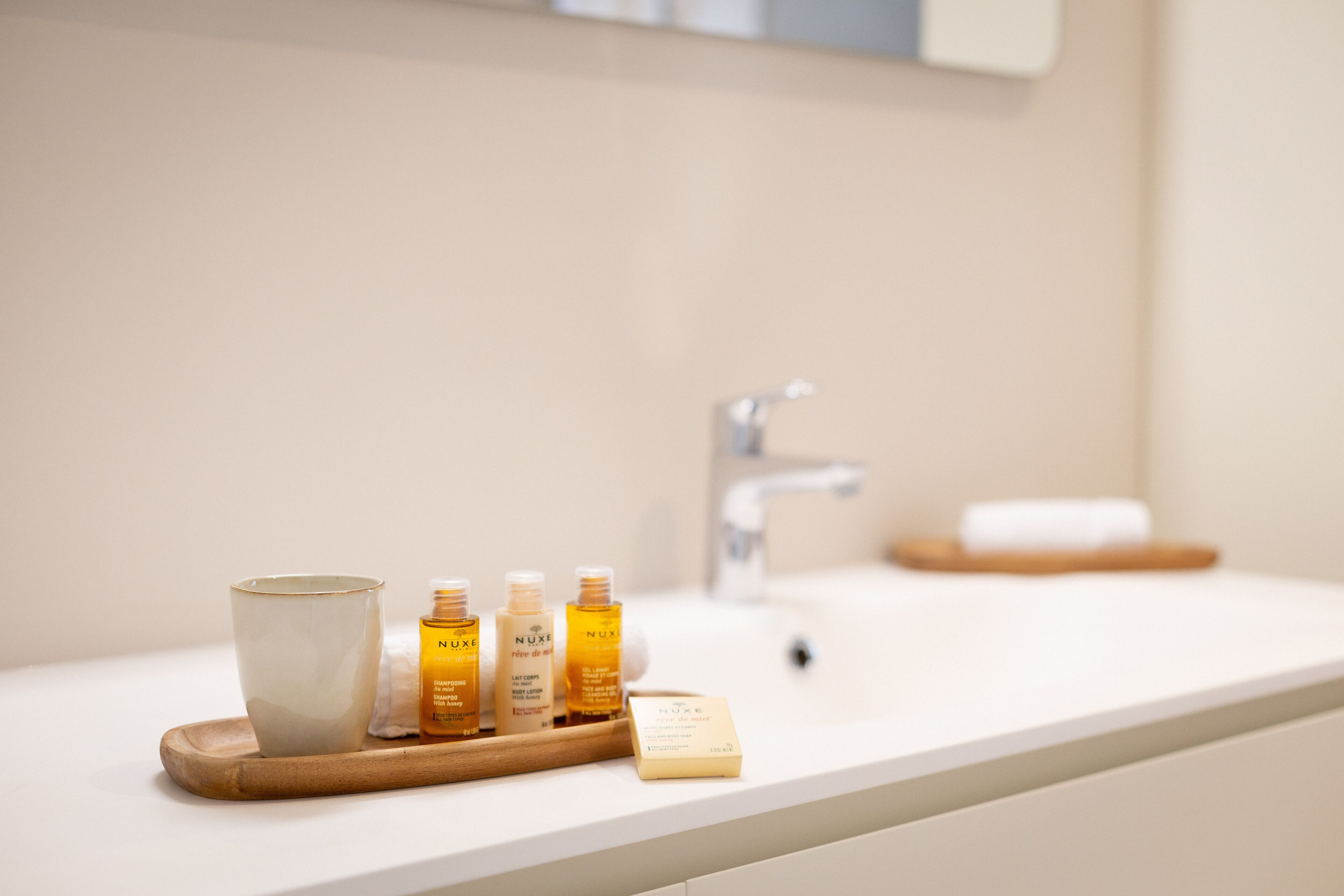 A bathroom counter with a white sink, a wooden tray holding a ceramic mug and three Nuxe skincare bottles, and a soap bar.