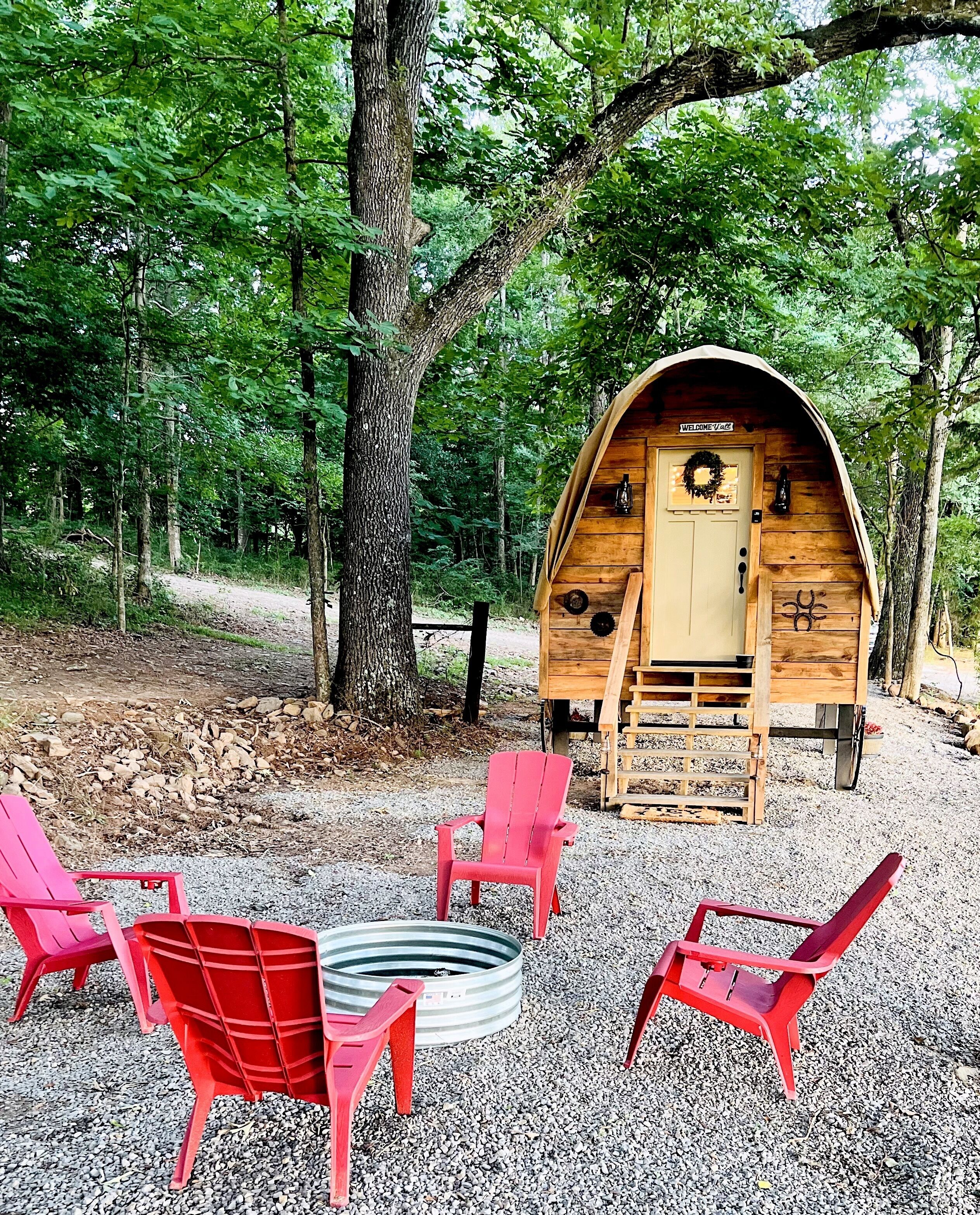 A rustic wooden cabin with a welcoming sign, surrounded by a gravel area with red chairs.