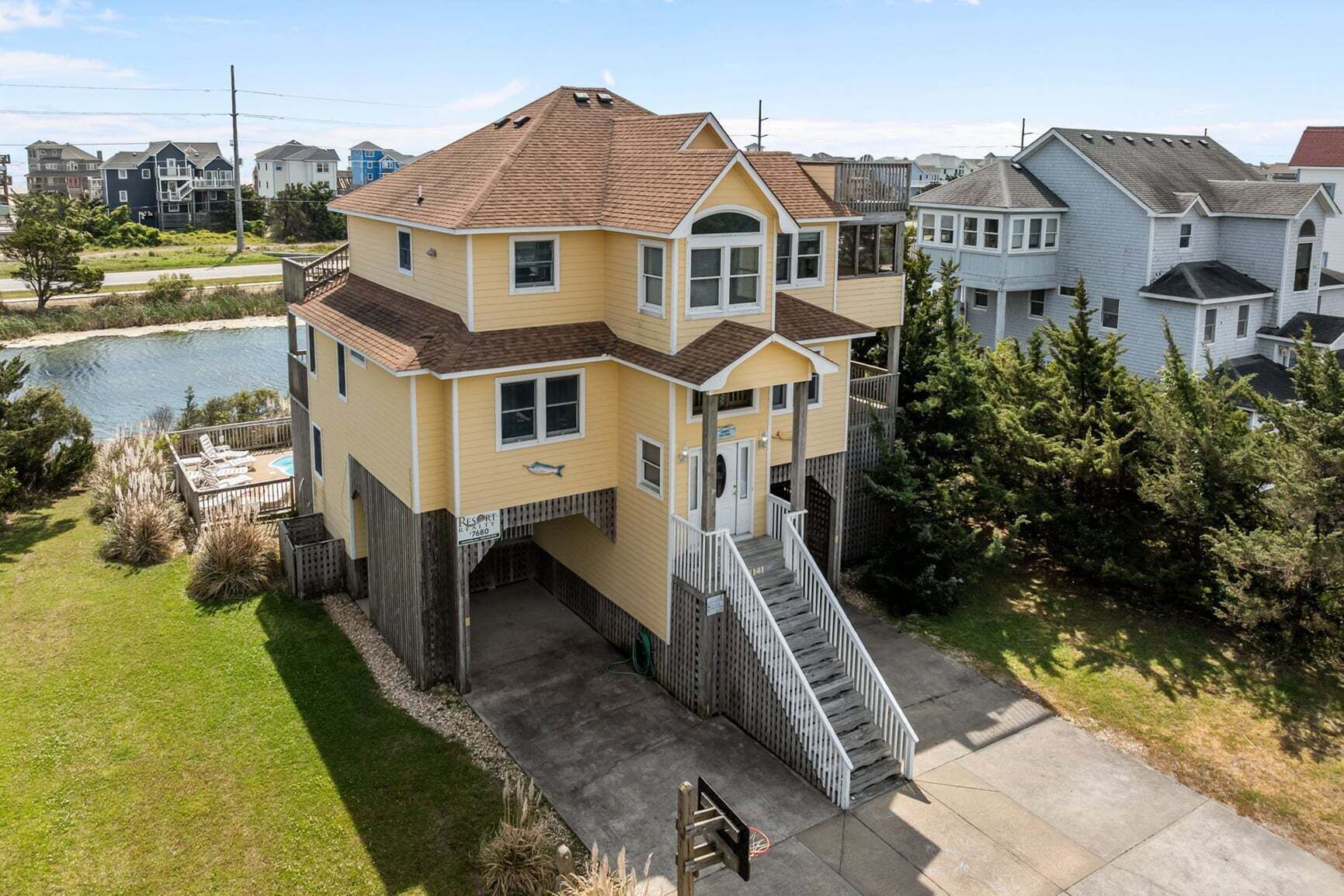 A two-story house with a front deck and a small pool, surrounded by other houses and a body of water.