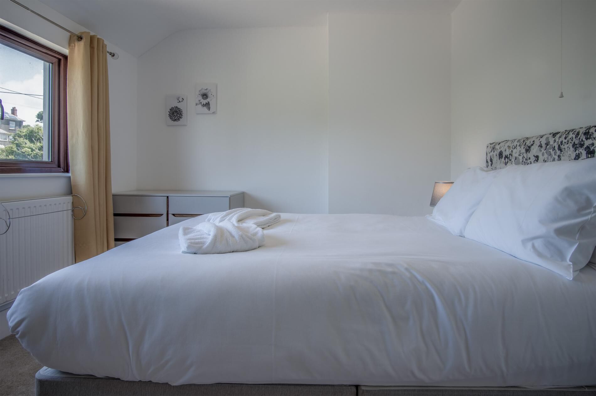 Una cama bien hecha con sábanas blancas, una mesita de noche pequeña y una ventana con cortinas.