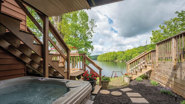 A wooden deck with a hot tub, stone steps, and a view of a lake surrounded by trees. - Cozy Dog Friendly Retreat w/ Hot Tub & Views (Mchenry)