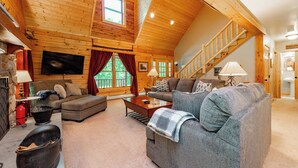 A cozy log cabin interior with a living room featuring a grey sofa, a wooden coffee table, and a fireplace. A staircase leads to an upper level, and a television is mounted on the wall. - Cozy Dog Friendly Cabin Near State Park w/ Hot Tub (Oakland)