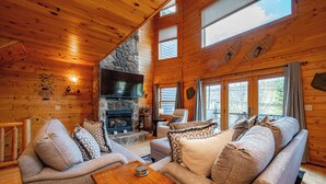 A cozy log cabin interior with a fireplace, comfortable seating, and large windows.