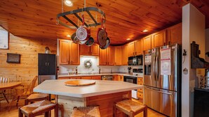 A kitchen with wooden cabinets, a large island with stools, stainless steel appliances, and a hanging pot rack.
