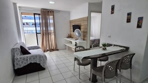 A modern living room with a dining area, white tiled floor, a sofa with a patterned cover, a table with chairs, a ceiling fan, and a view through a large window with curtains. - Apartamento Beira Mar Pajuçara Maceió (Maceió)