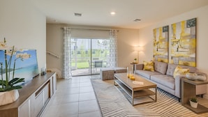 A modern living room with a sofa, coffee table, and TV. There is a large window with curtains, a rug, and artwork on the wall.