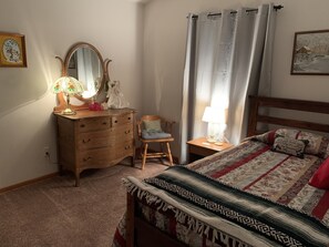 Ein Schlafzimmer mit einem hölzernen Schminktisch, einem Sessel, einem Bett mit gemustertem Überwurf, einem Nachttisch mit Lampe, einem Spiegel und einem Fenster mit Vorhängen.