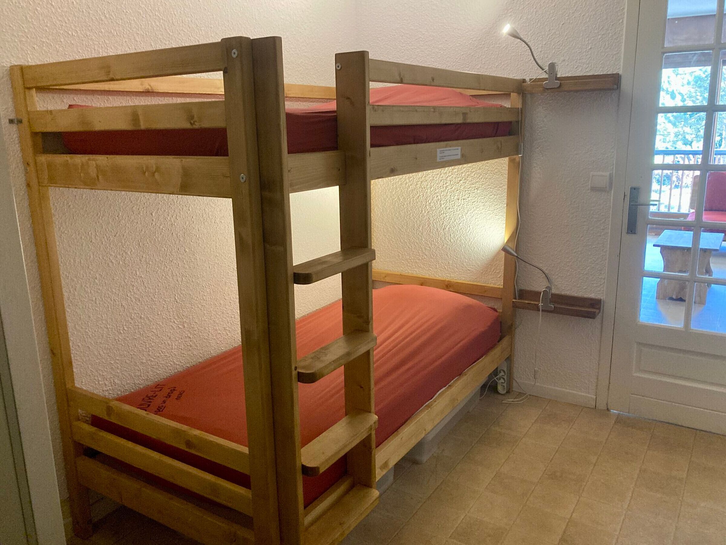 A bunk bed with a red mattress in a room with white walls and a door leading to an outdoor area.