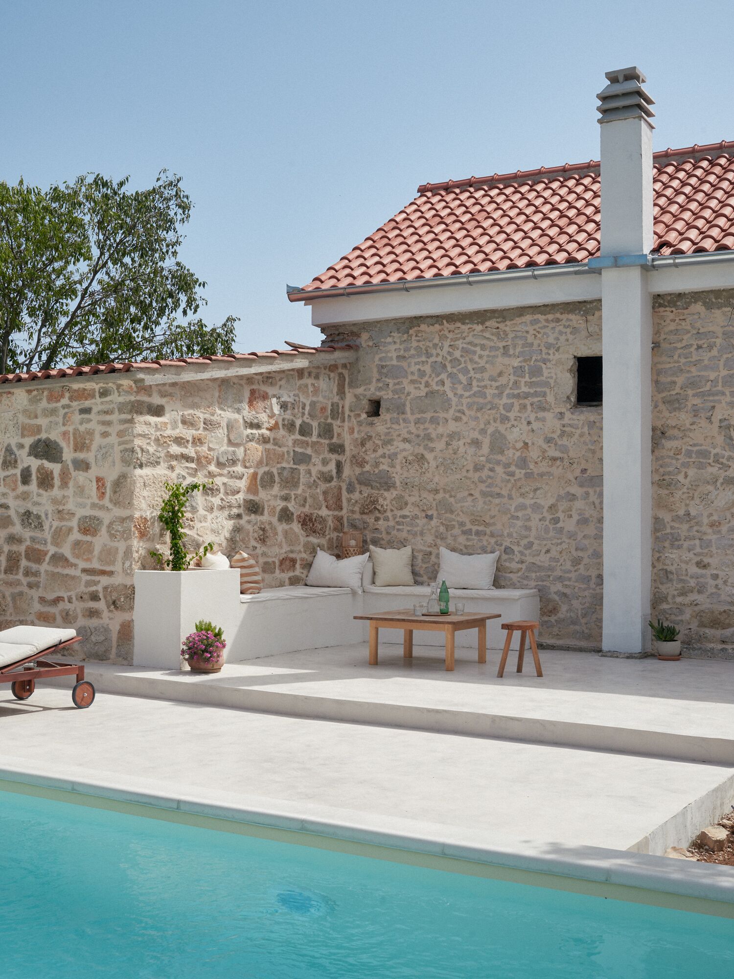 A stone building with a tiled roof, a pool, a wooden table, and a white sofa with cushions.