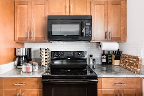 A kitchen with wooden cabinets, a black microwave, a stove, and various kitchen items on the countertop.