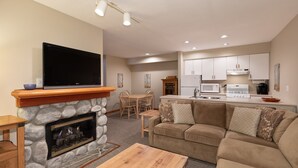 A living room with a stone fireplace, a flat-screen TV, a wooden coffee table, a beige sofa, and a kitchen area with white cabinets and appliances. - Lake Placid Lodge by Whistler Premier (Whistler)