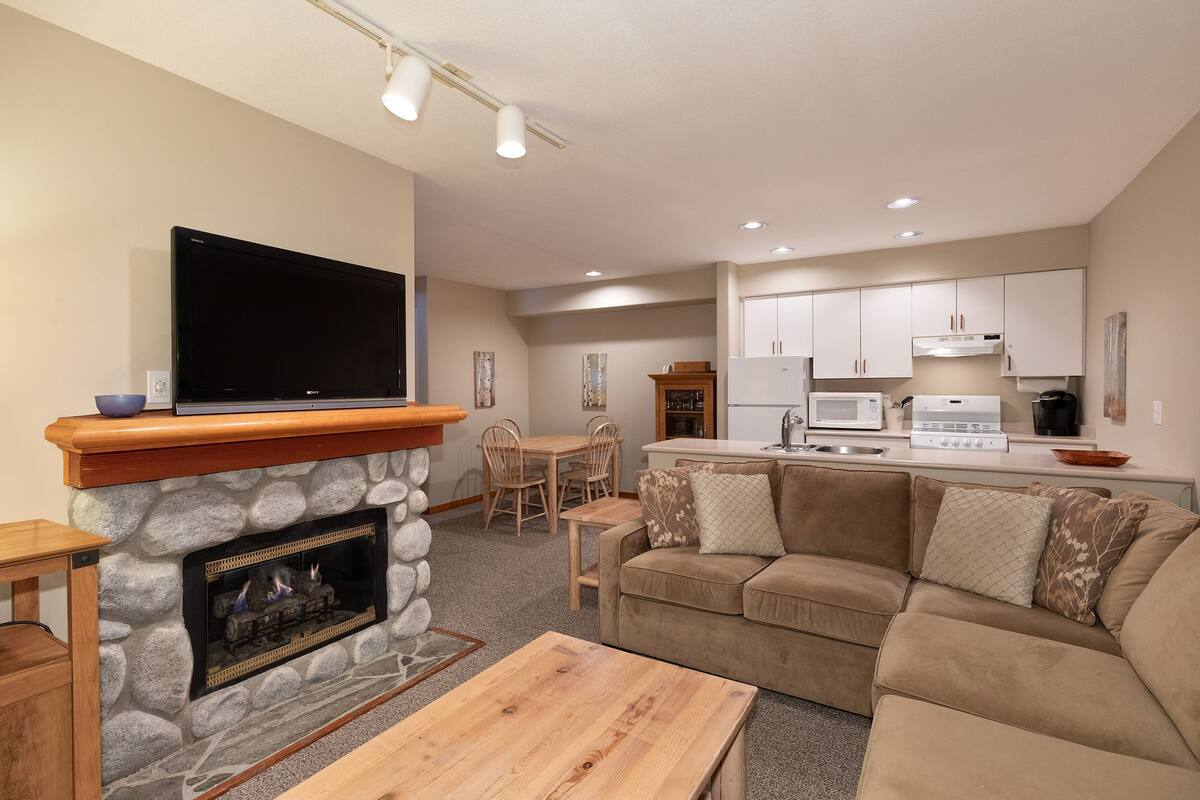 A living room with a stone fireplace, a flat-screen TV, a wooden coffee table, a beige sofa, and a kitchen area with white cabinets and appliances.
