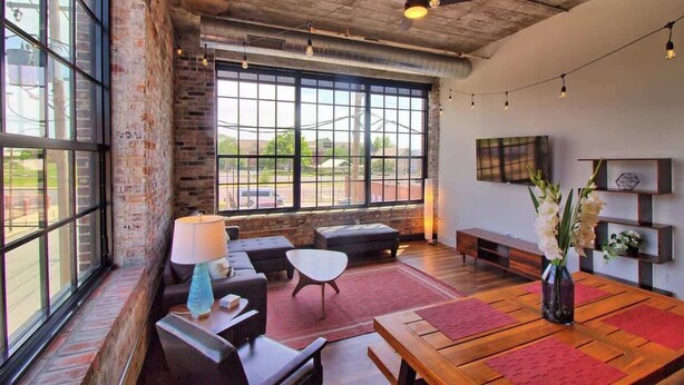 A spacious room with large windows, a brick wall, a wooden table, and a sofa. - Gorgeous Ultramodern 2BR Corner Loft + Workspace (Saint Louis)