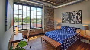 A bedroom with a large window, a bed, a desk, and a bench. - Gorgeous Ultramodern 2BR Corner Loft + Workspace (Saint Louis)