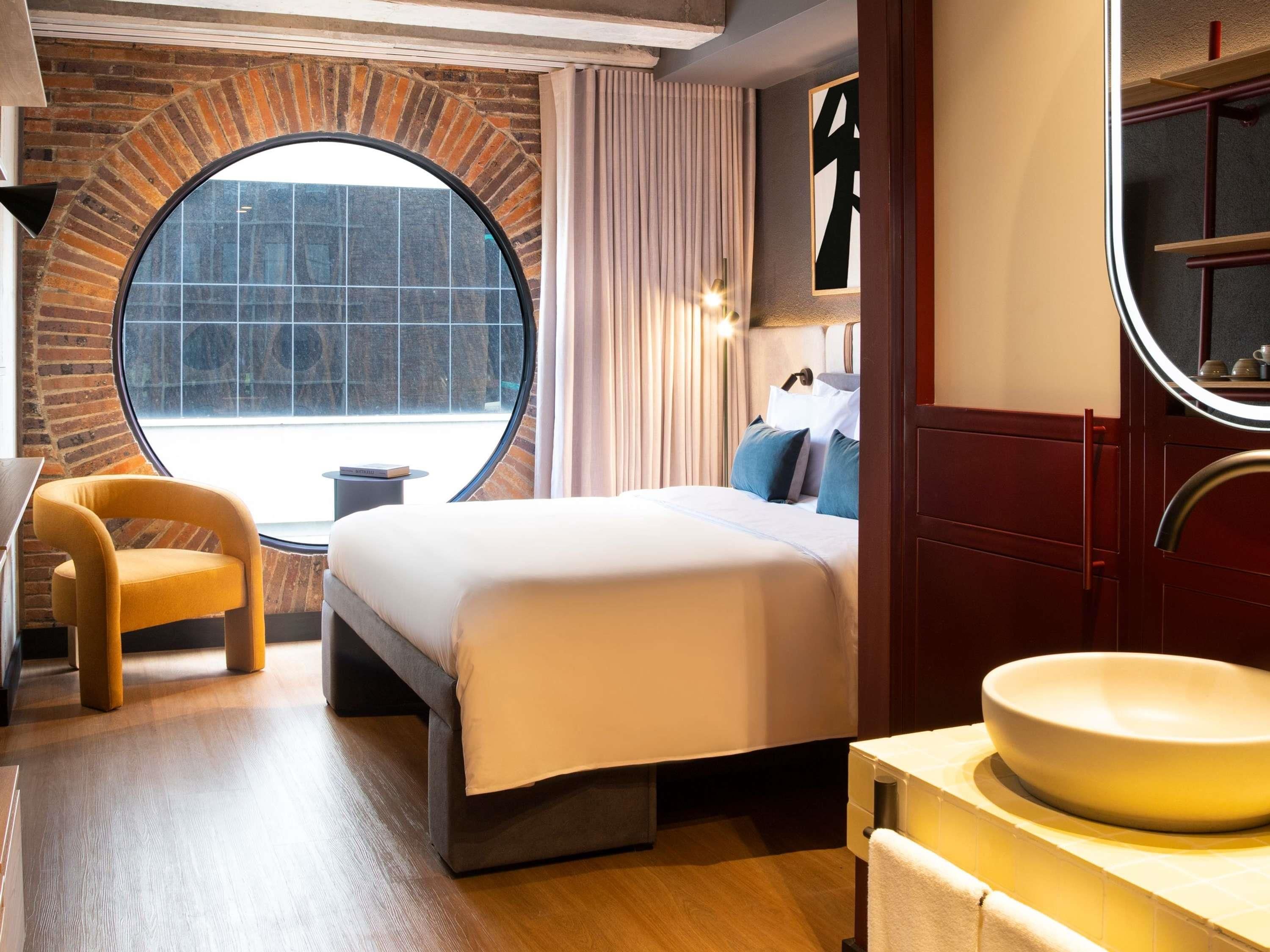 Une chambre d’hôtel avec une fenêtre ronde, un lit, une chaise, un miroir rond et un lavabo.