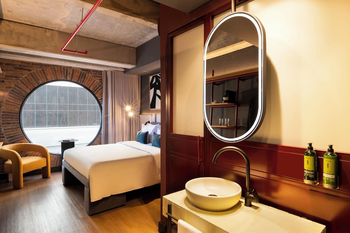 Una habitación de hotel moderna con un amplio ventanal circular, una cama con ropa blanca, un espejo redondo, un tocador de madera con lavamanos y dos botellas verdes en la pared.