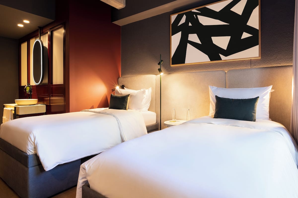 Habitación de hotel con dos camas, un espejo grande, una obra de arte en la pared y una mesita de noche con una lámpara.
