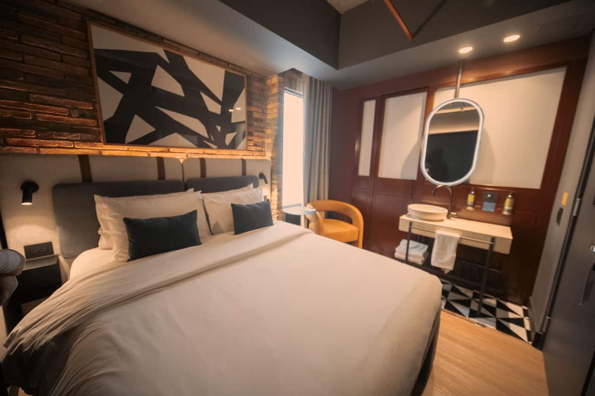 Una habitación de hotel moderna con una cama grande, cabecero de madera, baño con espejo y piso a cuadros.