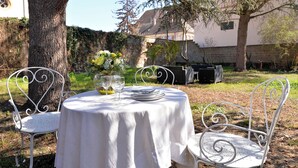 Une table et des chaises en métal blanc sont disposées à l’extérieur, recouvertes d’une nappe blanche, et entourées d’arbres. Un bâtiment se trouve en arrière-plan.