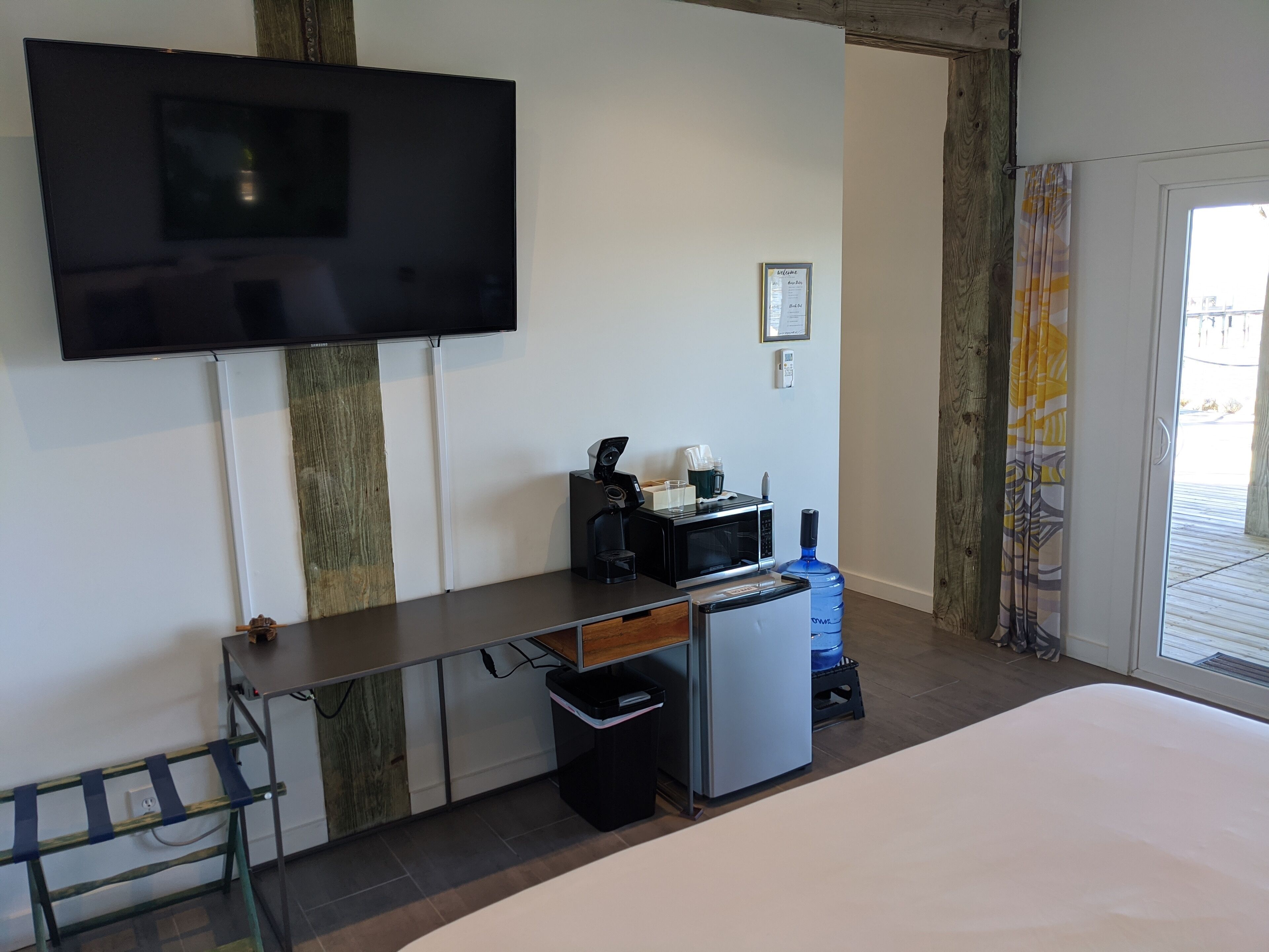Een hotelkamer met een flatscreen televisie aan de muur, een kleine kitchenette met een magnetron en koffiezetapparaat, een bed met een wit matras, en uitzicht op het omliggende landschap door een glazen deur.