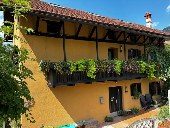 Un bâtiment jaune avec un toit recouvert de tuiles rouges, des fenêtres à encadrement noir et un balcon orné de plantes suspendues.
